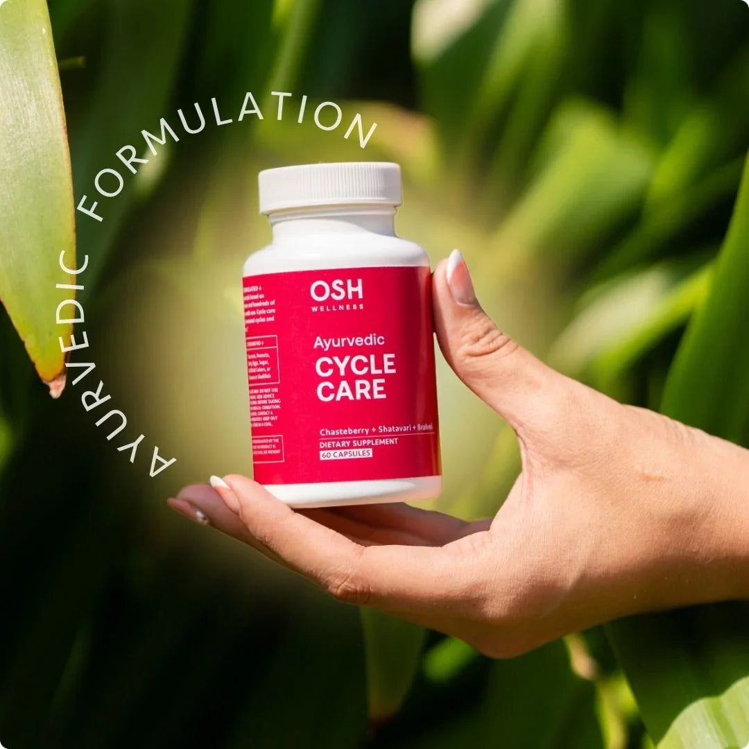A hand holds a bottle of OSH Wellness Ayurvedic Cycle Care dietary supplement in front of lush green foliage.