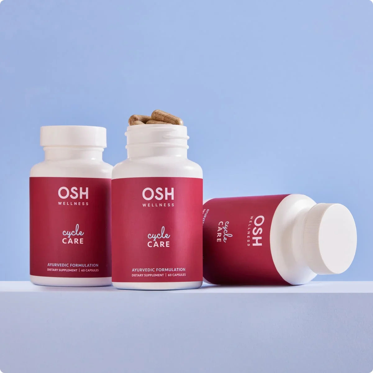 Three bottles of "OSH Wellness Cycle Care" supplements on a white platform against a blue background.