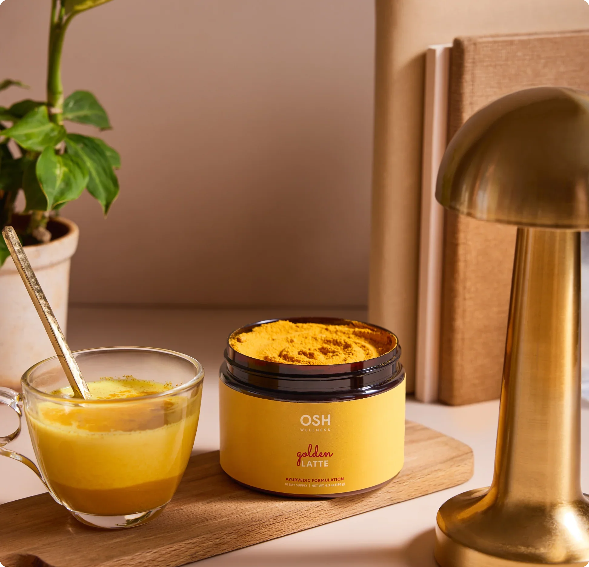 A jar of OSH golden latte powder mix next to a cup of golden latte, a lamp, and a plant.